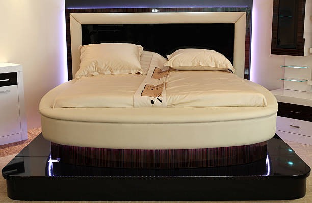 Luxurious round ottoman sofa bed with cream-colored sheets and black wooden base.