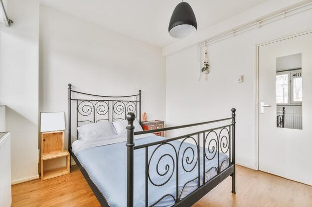 A minimalist bedroom with a small double metal bed frame featuring swirling designs, light blue bedding, a wooden bedside table, and a black pendant lamp.