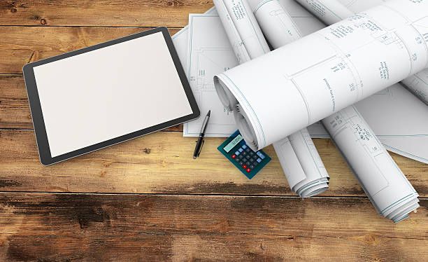 Wooden table with blueprints, a digital tablet, a black pen, and a calculator. Blueprints are rolled; tablet screen is blank.