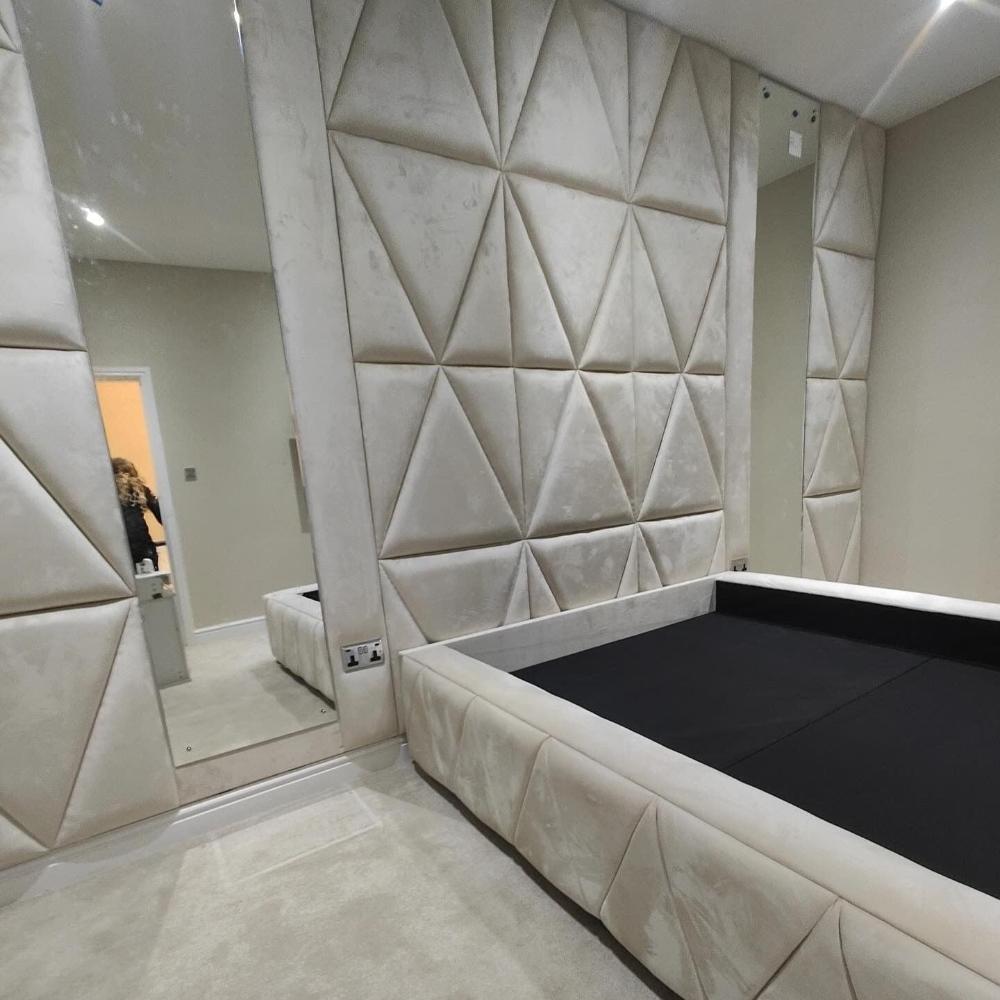 The image showcases a modern bedroom interior with distinctive wall and bed designs. The room features a large wall covered in padded wall panels with a triangular pattern, creating a textured, 3D effect. These panels are covered in a soft, light beige fabric that adds a sense of luxury. The bed frame, also covered in matching fabric, has a simple rectangular shape with an open top displaying a black surface.