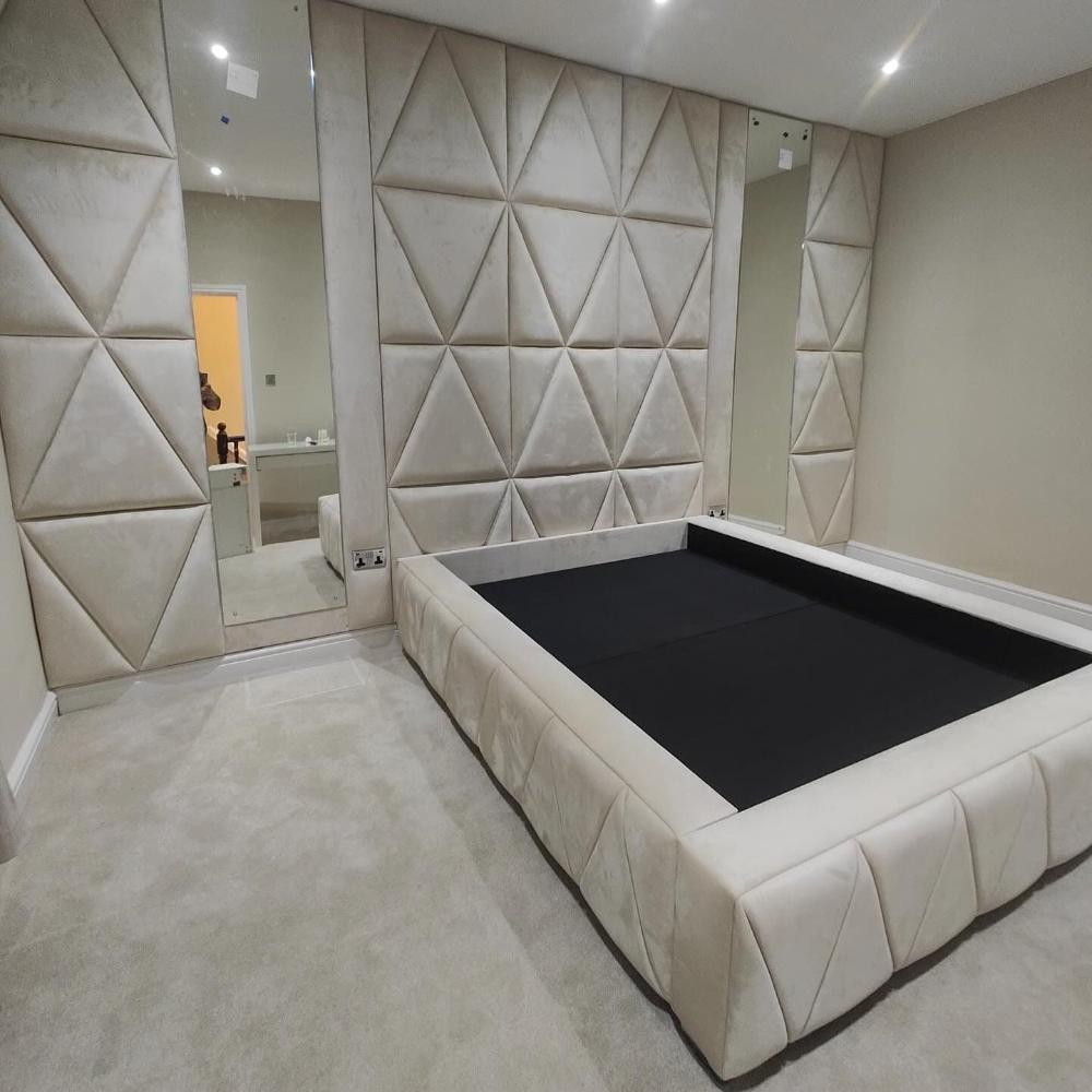 The image depicts a modern, minimalist bedroom with a distinctive design. The walls are covered in light beige, padded wall panels featuring a pattern of large, embossed triangular shapes. The flooring is covered in a matching light beige carpet. A large mirror is positioned on the left side of the room, reflecting part of the wall and an open doorway which reveals a different room with a person partially visible.