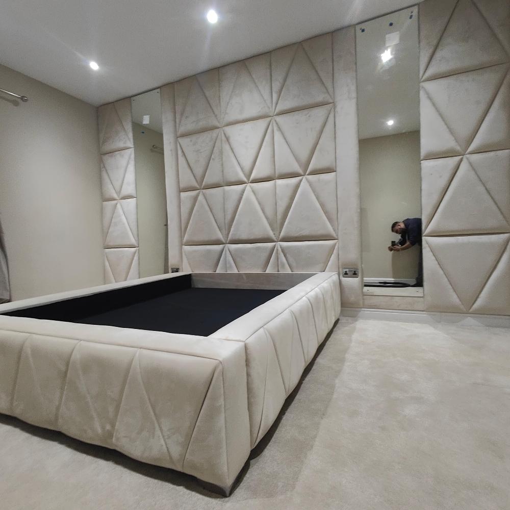 The image depicts a modern, elegantly designed room with a neutral beige color scheme. Central to the room is a low, rectangular bed frame upholstered in soft, light beige fabric with subtle geometric stitching. The bed is positioned on a plush, light-colored carpet. The walls are adorned with decorative wall panels featuring triangular patterns covered in matching fabric, adding texture and depth to the space. A tall, narrow mirror is integrated into the wall paneling to the right of the bed.