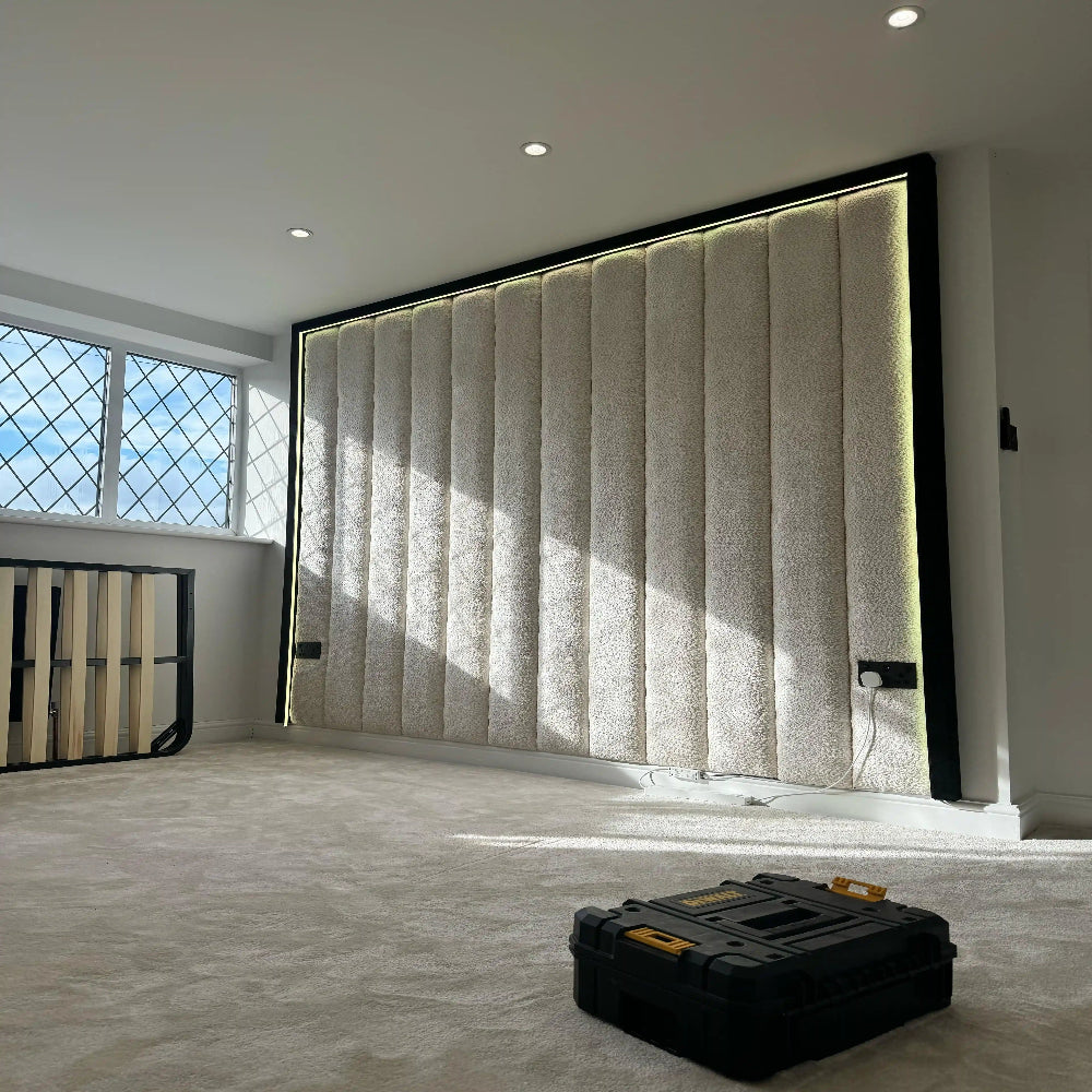 Room with a large fabric-paneled wall, black framings, soft lighting, window, and a toolbox on the carpeted floor.