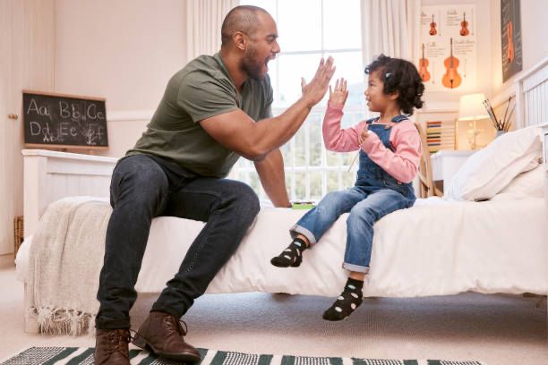 A smiling adult and child sit on a bed giving a high-five in a bright room with a chalkboard and musical instrument posters.