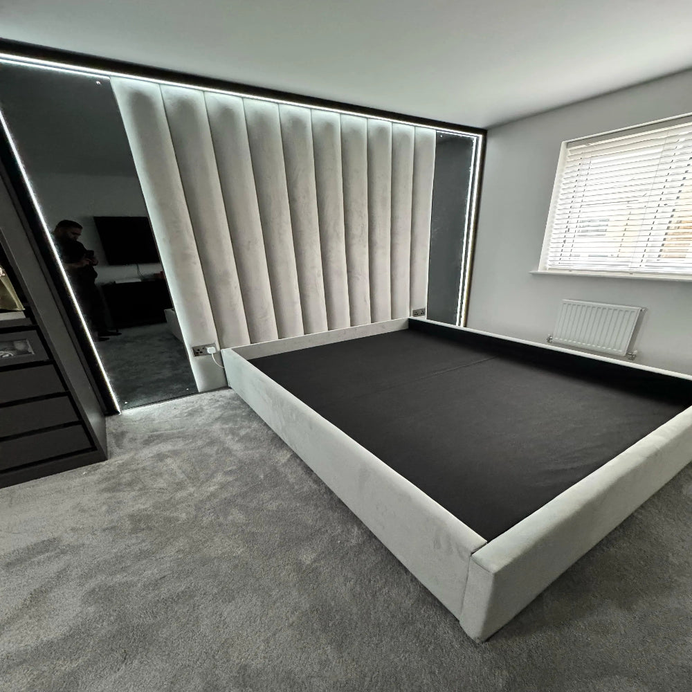 Modern luxury grey upholstered bed frame with tall panelled headboard, LED lighting and soft carpeted flooring.