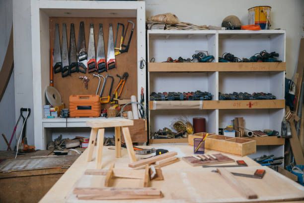 A cluttered woodworking bench with saws, tools, and wooden pieces in a neat woodshop with tools and equipment organized on shelves.