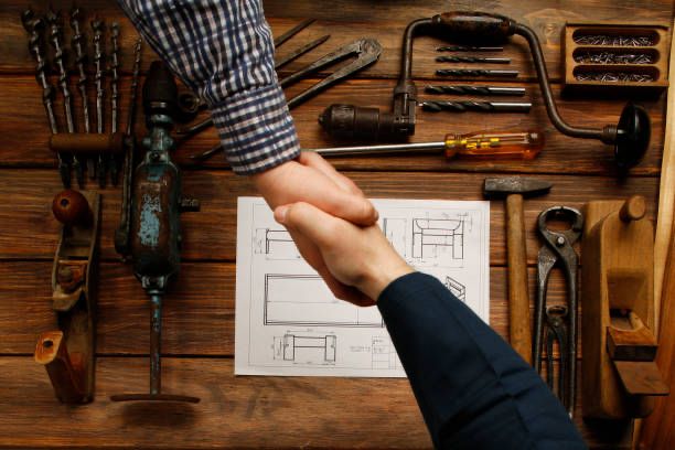 Two people shaking hands over a table with woodworking tools and a blueprint, symbolizing a collaborative agreement in craftsmanship.
