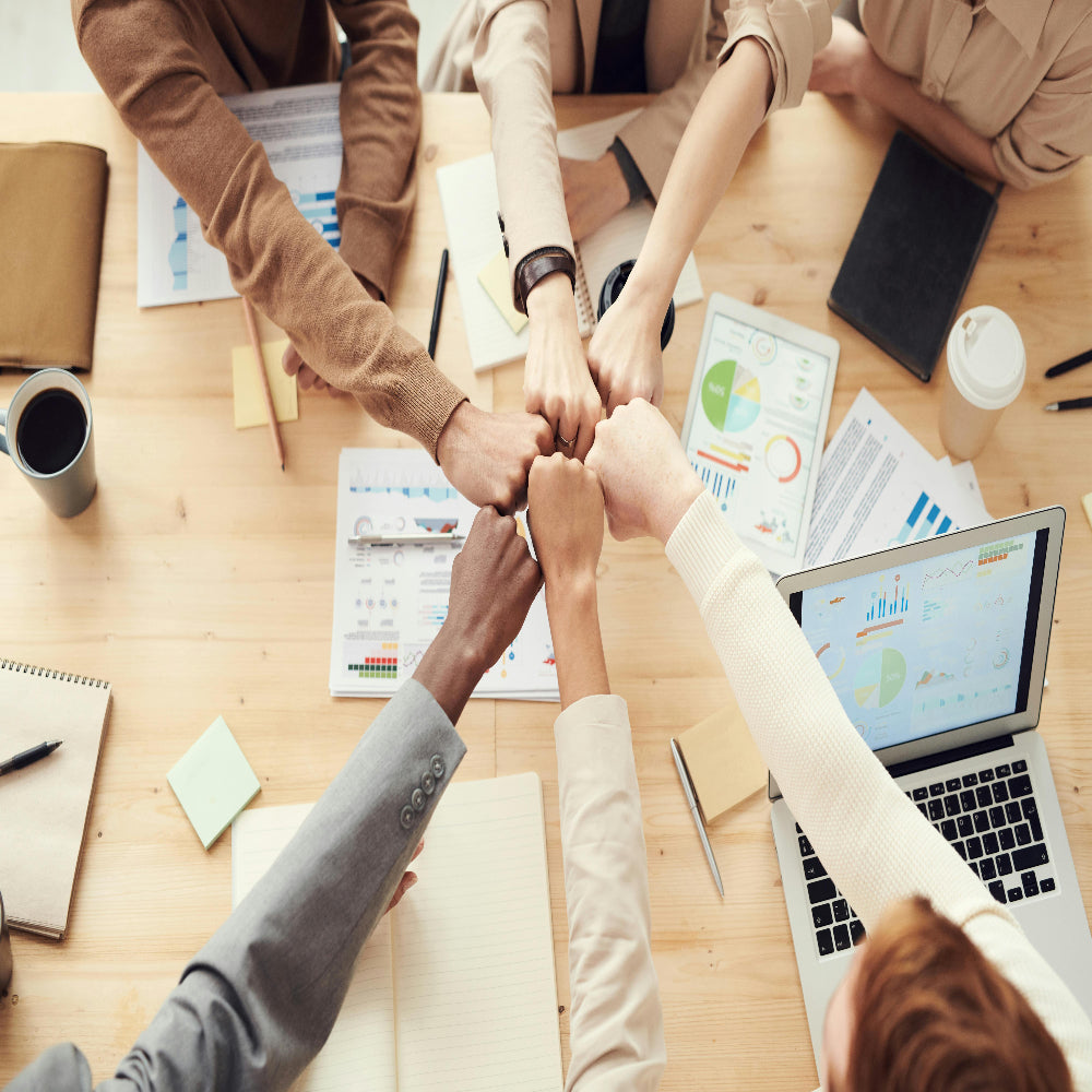 Top view of six people around a table performing a fist bump over charts, graphs, a laptop, notebooks, and a coffee cup, symbolizing teamwork.