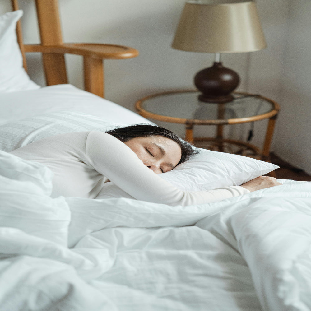 Person sleeping on a bed with white bedding, head resting on a pillow, near a wooden headboard and a bedside table with a lamp.