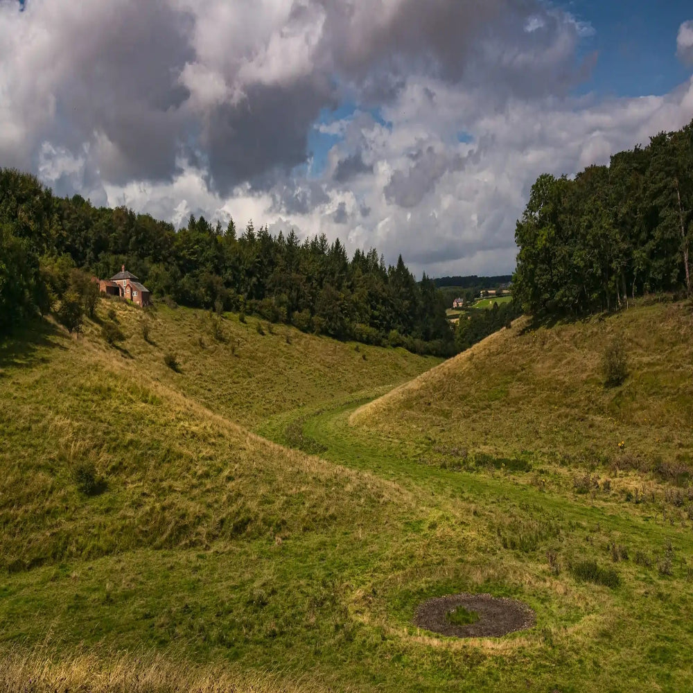 A green valley landscape with forested slopes, a cottage, cloudy sky, and a circular patch of grass in the foreground.