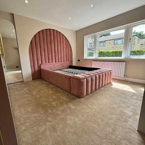 A blush pink upholstered twin bed size frame with an arched headboard in a beige bedroom, next to a large window.